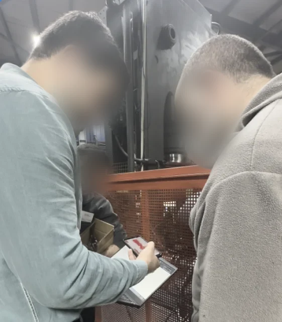 Three people looking at a phone and a tablet beside industrial machinery during a field visit, faces blurred